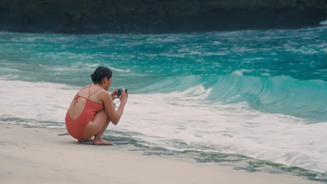 Anonymous girl sitting on green reef at sea comb and shooting splashes of crashing waves of blue ocean in summer using cell phone and waterproof case. Woman filming breaking sea water on wild seaside.