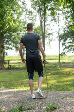 Back view photo of young man walking in park and holding jump rope.