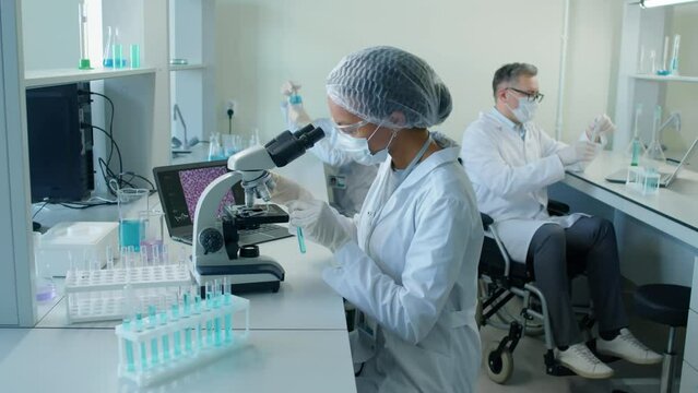 Arc shot of Biracial young woman in white lab coat, goggles and mask sitting at desk looking at probes in microscope in laboratory at daytime