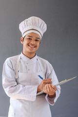 Young Asian male chef in white apron and toque writing in a clipboard doing inventory isolated over grey background.