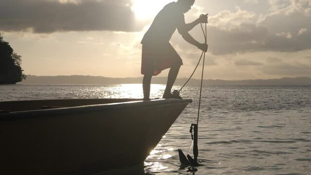 Sailing travel, anchor and boat of a fisherman on the ocean water on holiday outdoor. Cruise, sailor and man working on pulling a rope with transportation and adventure by the sea and beach at sunset