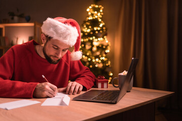Fototapeta na wymiar Men wearing Santa hat writing greeting cards at home office during christmas