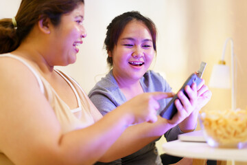 Happy cheerful Asian oversize - fat women sitting on the bed in bedroom in the night enjoy eating a snack or french fry and making an online shopping on a smartphone or laptop together in the night.