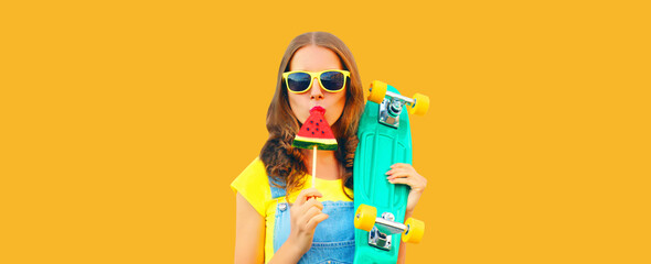 Summer portrait of young woman 20s with skateboard eating ice cream shaped slice of watermelon or sweet lollipop on orange background © rohappy