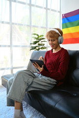 Attractive young gay man wearing headphone using digital tablet, surfing social media, watching movies while relaxing on couch at home