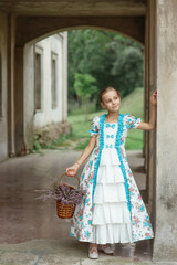 Young pretty girl in vintage elegant old-fashioned fluffy dress standing with basket of heather flowers in arch of old mansion in countryside, romantic summer outdoor story  in provence retro style