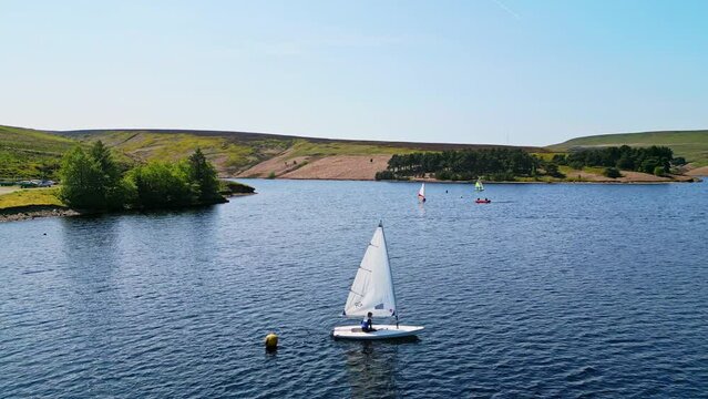Bright sunlight illuminates the Winscar reservoir in Yorkshire as sailing club members engage in a thrilling boat race with their white-sailed boats on the calm blue lake.