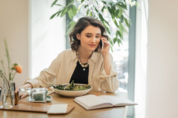 positive woman in stylish casual clothes looking at notebook during conversation on mobile phone near fresh vegetable salad, glasses and cup of aromatic coffee in hotel lobby, work-life integration