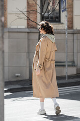 Obraz premium full length of young and trendy woman in grey hoodie, beige trench coat, white sneakers and dark sunglasses looking away and crossing in European city, street photography, urban fashion