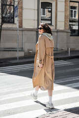 Fototapeta premium full length of fashionable woman in grey hoodie, beige trench coat and white sneakers crossing road and looking away in European city, travel lifestyle, urban fashion
