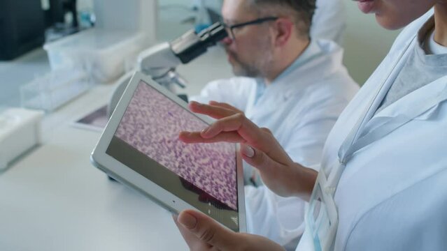 Focus on hands of unrecognizable woman in medical gown holding tablet studying picture of cells in laboratory at daytimeFocus on hands of unrecognizable woman in medical gown holding tablet studying p