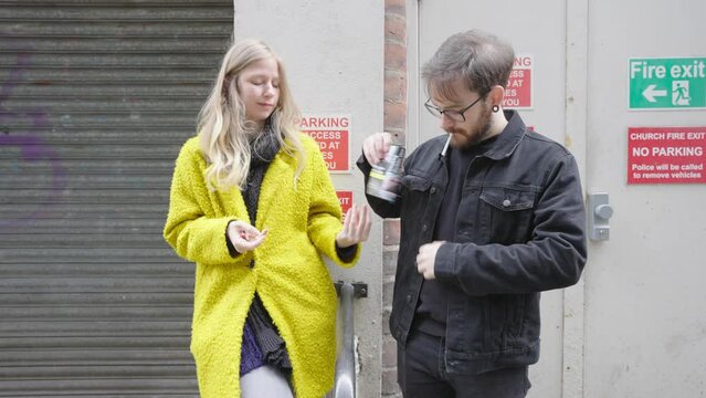 Female in yellow jacket and male in black jacket standing outside smoking