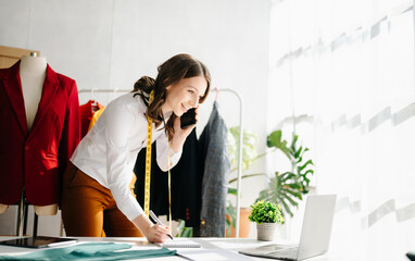 Fashion designer woman talking smart phone and using laptop with digital tablet computer in modern studio the clothes hanging on the racks .