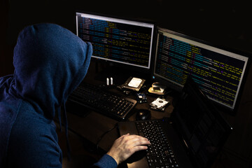 A hacker in a blue hoodie on a dark background in front of monitors that show the source code. A cyber criminal hacks the program.