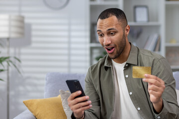 Shocked and happy young hispanic man using phone and credit card at home on sofa. Made a successful...