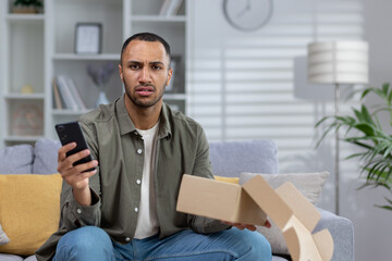Worried hispanic man sitting on sofa at home and holding parcel and phone. Looking at the camera...