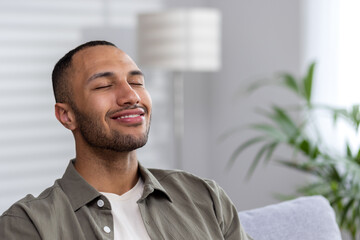 Portrait of the face of a smiling young Latin American man thoughtfully sitting at home on the sofa with closed eyes, resting, dreaming