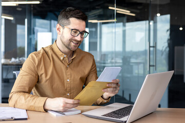 A young man businessman sits smiling in the office at the table and holds in his hands and reads documents, a contract, a successful deal that he received by mail