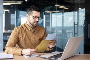 A serious and worried young businessman working in the office holds a letter, documents. got bad news. Saddenedly reads a message, a contract, an agreement