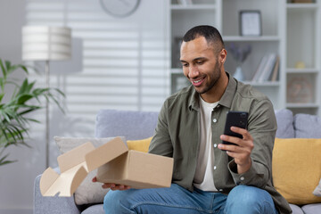 A happy young Latin American man is sitting on a sofa, holding a phone in his hand and is happy with a received gift, a parcel that was delivered to his home