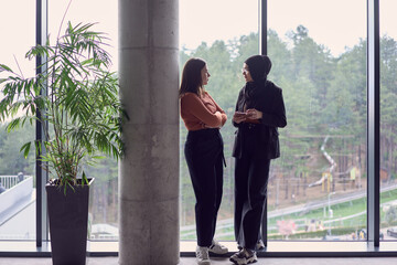 In a modern office a pair of colleagues, one wearing a hijab, engage in a lively discussion, sharing their business ideas and fostering a diverse and inclusive work environment.