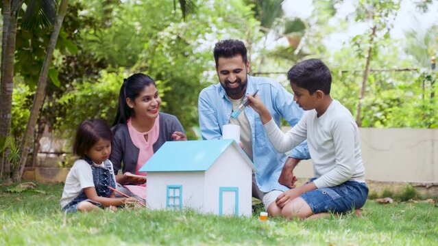 Happy indian parents helping kids to do painting on toy house at park - concept of Family art project, weekend holidays and Parental guidance.