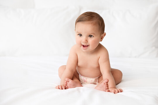 Baby girl wearing cloth diaper sitting on white bed leaning on hands with funny expression