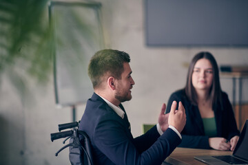 A wheelchair bound businessman confidently leads a business meeting in a large, modern office, exemplifying inclusive leadership, effective communication, and the power of diversity in driving success