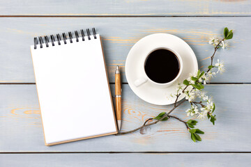 Morning coffee cup. Composition with opened blank notepad and pen, cup of black coffee, branch of white cherry blossoms on blue wooden table. Festive office desktop concept.