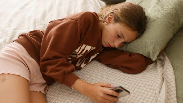 Sad depressed calm teenage girl laying on bed and scrolling through social networks on her smartphone