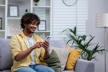 A young Hispanic man is sitting at home on the sofa in the living room, the man is holding a phone in his hands, using an online application on a smartphone, browsing the Internet and typing messages.