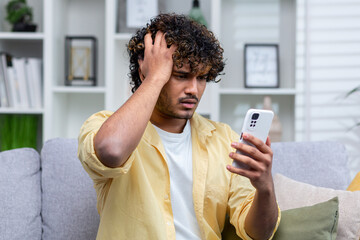 Latin american man sitting on sofa at home in living room, man upset and depressed reading bad news...