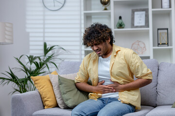 Young man sick at home sitting on sofa, Latin American has severe stomach pain and nausea, poisoned...