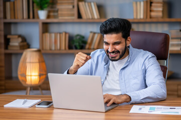 Overjoyed happy indian guy, lucky winner, sitting at desk watching virtual lottery results on laptop shaking hands. Eastern student got perfect exam grades, job proposal offer.