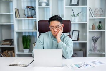 Obraz premium Bored sad young Asian guy sitting at desk using laptop, looking at pc screen, thinking about problems during online lesson or remote work at home office. Tired man leaning head on hand, watching news