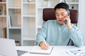 asian man freelancer or businessman working from home, sitting at table in front of modern laptop, having phone conversation, taking notes