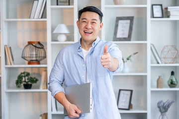 Happy asian man stands in a modern office, in a casual shirt, with a laptop in his hands, looks at the camera, smiles, shows thumbs up gesture