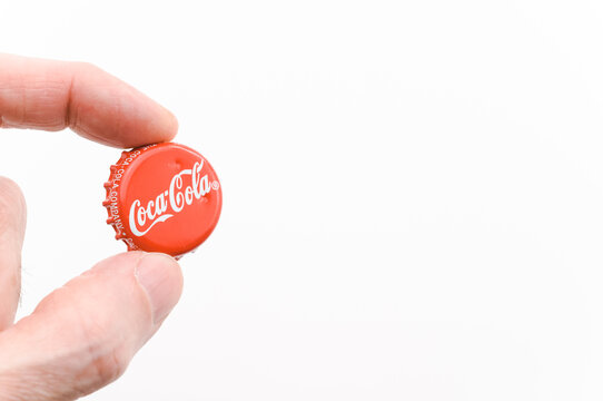 immagine editoriale illustrativa primo piano di dita che reggono  tappo classico in metallo a corona marchio Coca cola su superficie bianca