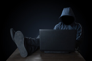 The hacker in the blue jacket threw his feet on the table. A cybercriminal hacks through a laptop.