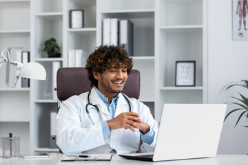 Young successful doctor working inside hospital office, man smiling and looking at laptop screen, hispanic man in medical gown is satisfied with patient's treatment achievement results.