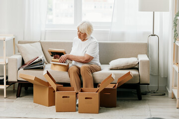 Fototapeta elderly woman sits on a sofa at home with boxes. collecting things with memories albums with photos and photo frames moving to a new place cleaning things and a happy smile. Lifestyle retirement. obraz