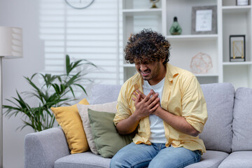 Sick man having severe chest pain and heart attack, hispanic young man sitting alone at home in...