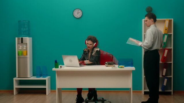 A woman freak sits in the office at a laptop in red headphones, listens to music, sings. An employee is standing nearby, looks at his colleague in a stunned way, leaves. Music application. Slow motion