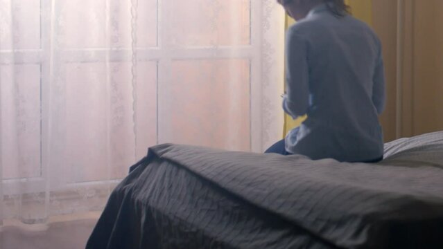 Lady in office clothes returns to room after work. Young woman sits on soft bed against window. Female looks out hotel window