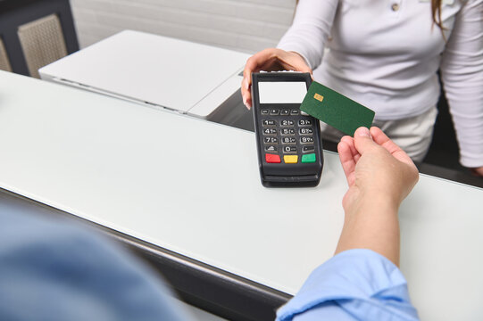 Hand of customer paying with contactless credit card with NFC technology. Female administrator with credit card reader machine at reception counter with female holding blank bank card. Selective focus
