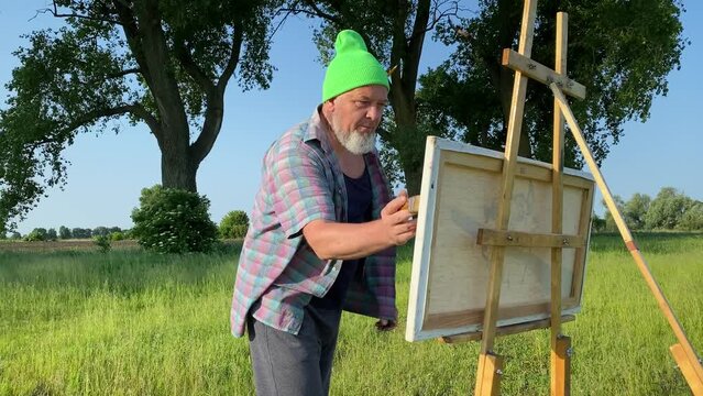 Dolly shot creative artisan applies oil paints uses paintbrush preparing canvas for painting. Beautiful scenic landscape meadow trees field on background. The Sun overflowing place of work in sunrise