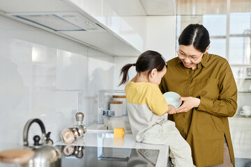 Happy Asian mom teaching to wash dish to her little daughter while they standing in the kitchen