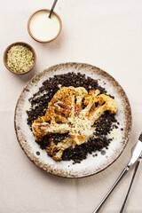 A white plate with brown rim with black beluga lentils and roasted cauliflower with paprika and black pepper seasoning, tahini dressing, on linen table cloth, a delicious high protein vegan meal