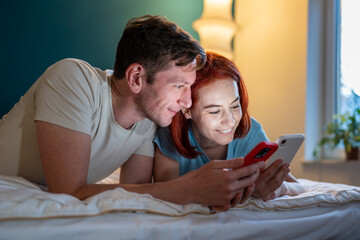 Fototapeta Couple looking phone while lying on bed. Girl showing boyfriend funny videos on phone. Happy husband and wife checking social media. Woman showing man new mobile phone app obraz