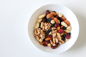 Top view of a white bowl of mixed nuts and dried fruits consisting of almonds, walnuts, hazelnuts, cashew nuts, raisins and cranberries, on right side of white table, copy space on left